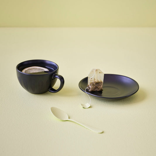 A yellow background showcases a cup with a spoon. The servingware features the iconic 'Lotus' pattern by Arne Clausen.