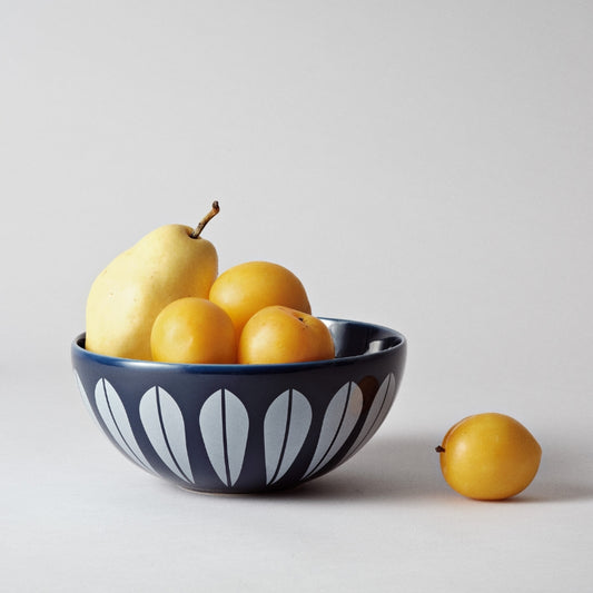 A bowl with a geometric lotus design filled with yellow fruits, including a pear and several round citrus fruits, on a plain background.
